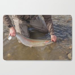 An angler unhooking a fly from a steelhead, rainbow trout, while it is still in the water, from a river river in British Columbia, Canada, in morning spring sun Cutting Board