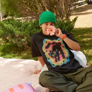 image of young man sitting on a picnic blanket outside wearing a green beanie and a funky cat tshirt.