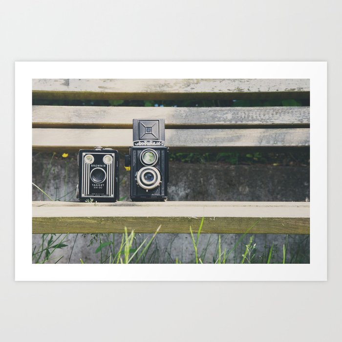 two vintage cameras sitting on a park bench ... Art Print