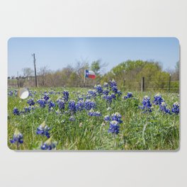 Bluebonnet flowers blooming by road with Texas flag in background Cutting Board