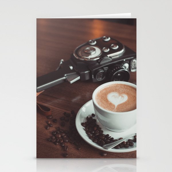 A cup of hot cappuccino placed on a table next to the old camera with lens and coffee beans Stationery Cards